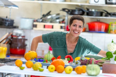 Mujer sonriente y relajada, con una pila de verduras orgánicas delante de ella con los utensilios de cocina en el fondo en foco.