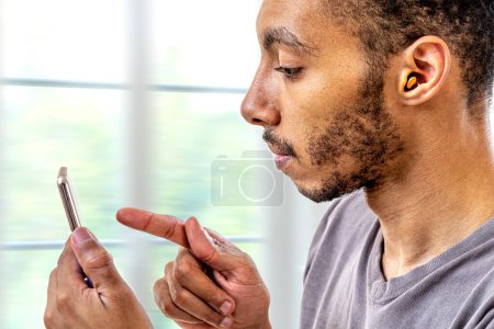 Junger Mann stellt mit dem Smartphone die Lautstärke des Hörgeräts ein.