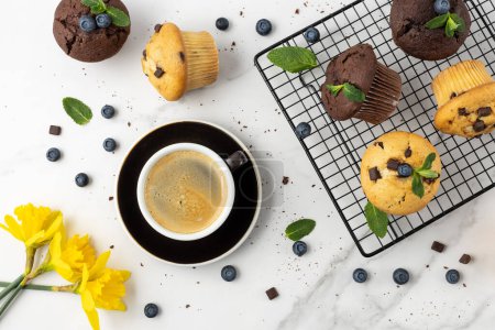 Photo for Cup of hot coffee, fresh baked muffins with chocolate chips, blueberry berries and mint leaves on white marble table background. Chocolate cupcake closeup as dessert for coffee break. Top view. - Royalty Free Image