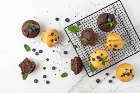 Photo for Fresh baked muffins with chocolate chips, blueberry berries and mint leaves on baking rack and on white marble table background. Chocolate  cupcake closeup as dessert for coffee break. Top view. - Royalty Free Image