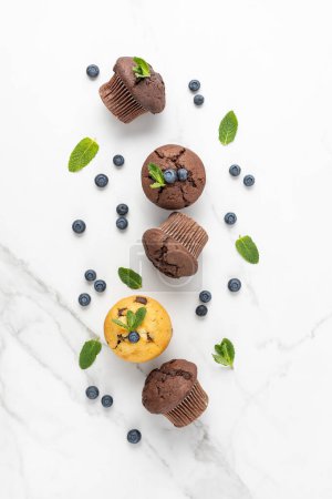 Photo for Fresh baked muffins with chocolate chips, blueberry berries and mint leaves on white marble table background. Chocolate and banana muffin or cupcake closeup as dessert for coffee break. Top view. - Royalty Free Image