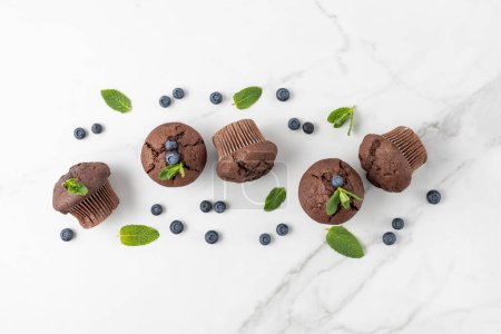 Photo for Fresh baked muffins with chocolate chips, blueberry berries and mint leaves on white marble table background. Chocolate cupcake closeup as dessert for coffee break. Top view. - Royalty Free Image