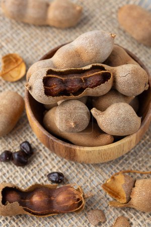 Ripe whole and shelled tamarind fruits with seeds in wooden bowl closeup on jute background. Healthy exotic food.