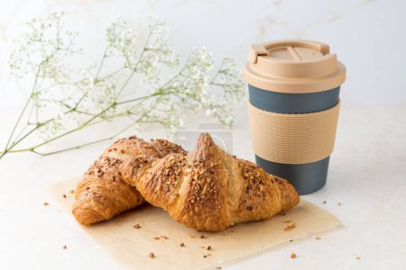 Fresh baked butter nougat crushed nuts breakfast croissant on paper with crumbs, coffee to go mug  on  light marble table background. Coffee break with pastry.