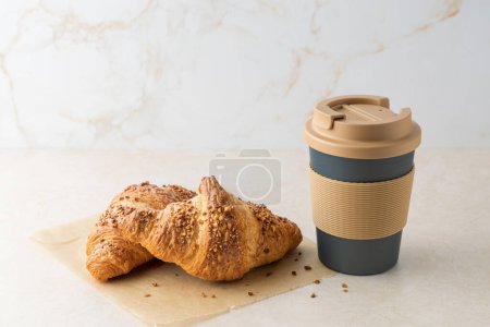 Fresh baked butter nougat crushed nuts breakfast croissant on paper with crumbs, coffee to go mug on light marble table background. Coffee break with pastry.