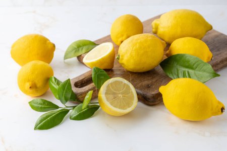 Fruits de citron jaune bio frais entiers et tranchés avec des feuilles d'agrumes vertes sur planche à découper en bois sur fond de table en marbre clair.