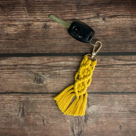 Blick von oben auf den handgefertigten Makramee-Schlüsselanhänger, der auf dem Autoschlüssel isoliert auf dem Holzhintergrund hängt.