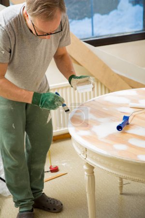 Middle-age man painting old wooden table with paint brush in white color. Furniture restoration, DIY repair, repair by oneself and home improvement. Recycling and sustainable lifestyle