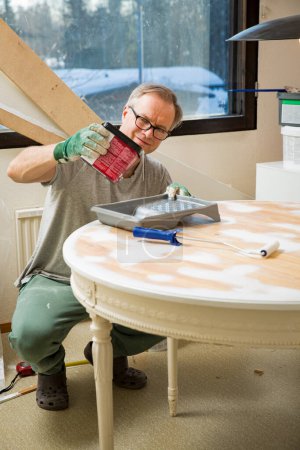 Middle-age man painting old wooden table with paint roller in white color. Furniture restoration, DIY repair, repair by oneself and home improvement. Recycling and sustainable lifestyle