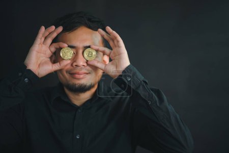 Bitcoin, digitales Geld. Porträt eines spielerisch aufgeregten bärtigen Mannes, der die Augen mit goldenen Bitcoins bedeckt und Vertrauen in die Kryptowährung hat