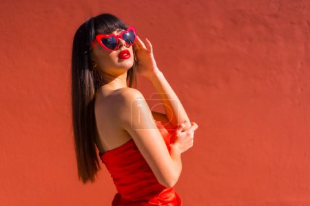 Téléchargez les photos : Brunette fille sur un fond rouge avec des lunettes de coeur et robe rouge serrée, Saint Valentin - en image libre de droit