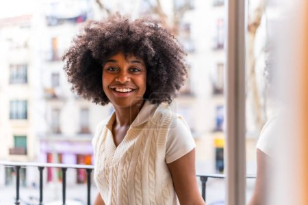 Foto de Retrato de una joven mujer étnica negra con cabello afro en un balcón en casa sonriendo, situación cotidiana, al atardecer mirando a la cámara - Imagen libre de derechos