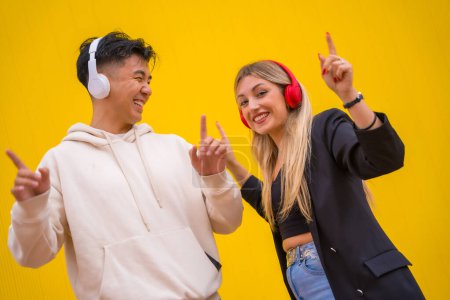 Multiethnic couple of Asian man and Caucasian woman on a yellow background, dancing bachata and salsa with headphones