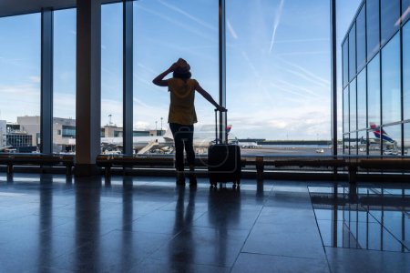 Téléchargez les photos : Passager femme avec bagages d'une valise dans l'aérogare regardant les avions par la fenêtre - en image libre de droit