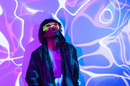 Retrato de estudio con luces de neón púrpura y azul de un hombre futurista afro mirando hacia arriba usando gafas de realidad aumentada