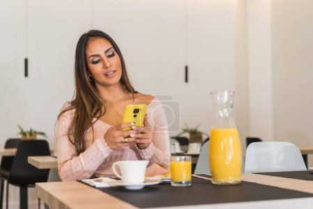 Téléchargez les photos : Beauté et femme d'affaires occasionnelle utilisant le téléphone tout en prenant le petit déjeuner dans un hôtel de luxe le matin - en image libre de droit