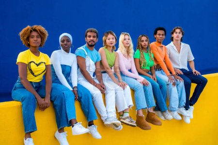 Téléchargez les photos : Groupe sérieux de jeunes multi-ethniques assis sur un mur coloré et regardant la caméra - en image libre de droit