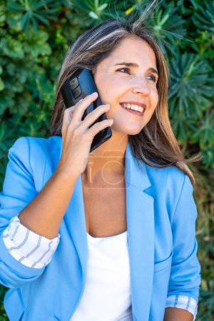 Photo for Vertical close-up portrait of a casual businesswoman talking to the phone in a green area - Royalty Free Image