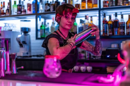 Téléchargez les photos : Jeune femme barman secouant un cocktail avec un shaker dans le comptoir d'un bar la nuit - en image libre de droit