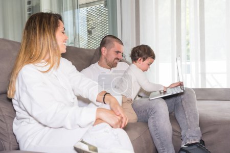 Téléchargez les photos : Bébé jouant avec le clavier d'un ordinateur portable assis sur le canapé avec ses parents - en image libre de droit