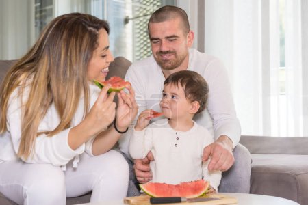 Téléchargez les photos : Nouvelles expériences pour un bébé garçon dégustant de la pastèque avec ses parents dans le salon - en image libre de droit