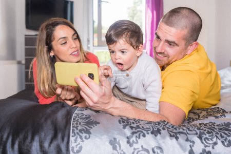Téléchargez les photos : Surpris bébé garçon regardant le mobile couché sur le lit avec ses parents - en image libre de droit