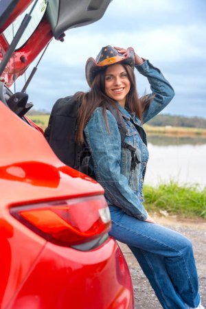 Téléchargez les photos : Photo verticale d'une jolie fille avec un chapeau de cow-boy assis sur une voiture rouge lors d'un voyage sur la route - en image libre de droit