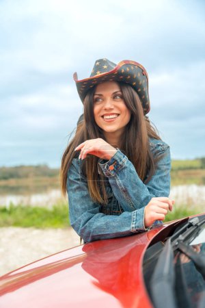 Téléchargez les photos : Portrait vertical d'une femme souriante avec un chapeau de cow-boy appuyé sur une voiture dans une route - en image libre de droit