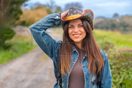 Téléchargez les photos : Femme rayonnante souriante avec chapeau de cow-boy marchant sur un chemin rural tout en voyageant seul - en image libre de droit