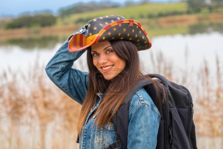 Téléchargez les photos : Femme sensuelle en chapeau de cow-boy souriant à la caméra à l'extérieur près d'un lac - en image libre de droit