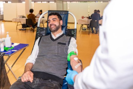 Man smiling sitting on chair next to an unrecognizable doctor in a blood donation pavilion