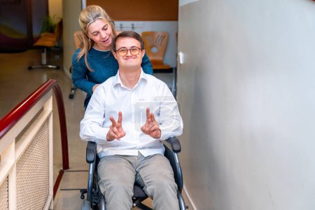 Téléchargez les photos : Travailleur de la santé poussant un homme heureux avec le syndrome du duvet en utilisant un fauteuil roulant alors qu'il gestuelle succès avec les doigts - en image libre de droit