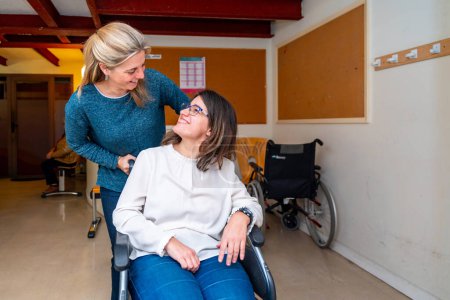 Téléchargez les photos : Aide soignante poussant le fauteuil roulant d'une femme handicapée dans un centre de jour pour les personnes ayant des besoins spéciaux - en image libre de droit