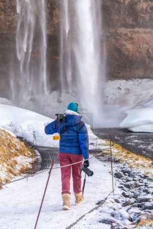 Téléchargez les photos : Photographe par derrière en hiver en Islande visitant la cascade Seljalandsfoss - en image libre de droit