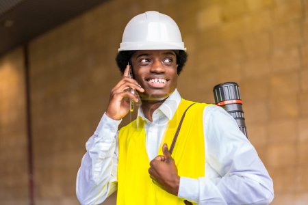 Porträt eines glücklichen jungen afrikanischen Architekten in Arbeitskleidung und Helm, der während eines Baustellenbesuchs mit dem Telefon spricht