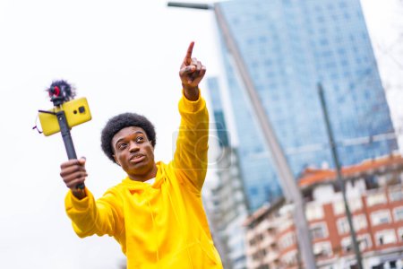 Afrikanischer junger Vlogger zeigt während eines Online-Videos mit einem Handy, das an Mikrofon und Stativ in der Stadt befestigt ist, nach vorne