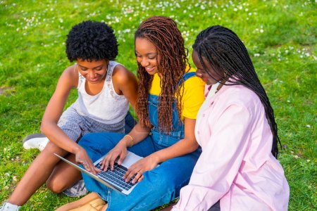 Téléchargez les photos : Vue du dessus de trois jeunes étudiantes africaines utilisant un ordinateur portable ensemble assis sur un parc - en image libre de droit