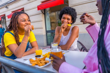 Trois jeunes amies africaines mangeant de la restauration rapide avec de la bière dans un restaurant en plein air