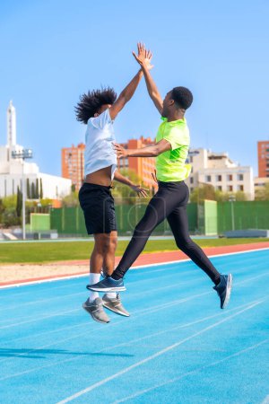 Vertikales Foto von zwei jungen afrikanisch-amerikanischen Freunden, die jubelnd auf einer Laufbahn springen und die Hände umklammern