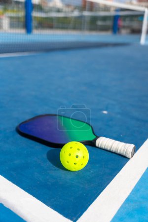 Foto vertical de una raqueta y una bola de pickleball colocadas en el suelo en una cancha azul