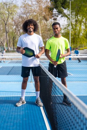 Retrato vertical de dos jóvenes jugadores afroamericanos de pickleball de pie mirando a la cámara sosteniendo equipo de pickleball