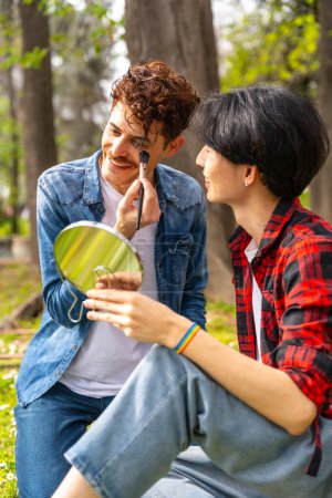 Photo for Vertical photo of two multi-ethnic gay men applying make up in a park - Royalty Free Image