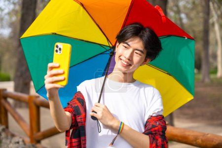 Photo for Chinese gay man taking a selfie with mobile phone while holding a rainbow colored umbrella standing in a park - Royalty Free Image