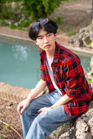 Photo for Vertical photo of an Asian gay man sitting next to urban stream of water in a park - Royalty Free Image