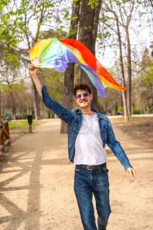 Vertikales Foto eines schwulen Mannes, der in einem Park einen regenbogenfarbenen Handfächer hebt und schwenkt