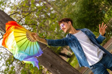 Photo for Dynamic shot of a happy casual gay male artist moving a rainbow fan dancing in a park - Royalty Free Image