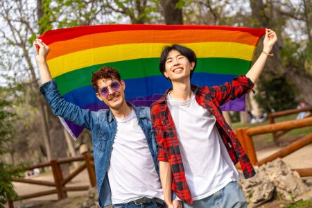 Photo for Portrait of a multi-ethnic gay couple raising a rainbow lgbt flag in a park - Royalty Free Image