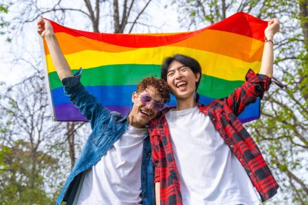 Photo for Low angle view portrait of a happy gay couple laughing while raising lgbt rainbow flag outdoors in a park - Royalty Free Image