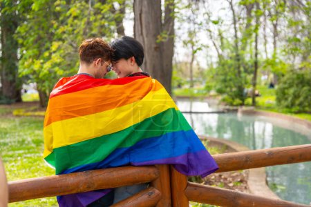 Rückansicht einer romantischen Szene eines multiethnischen homosexuellen Paares, das sich verliebt, in eine LGBT-Flagge gehüllt und in einem Park umarmt