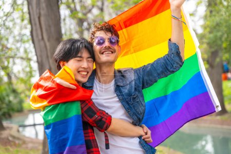 Photo for Happy multi-ethnic gay couple celebrating love waving lgbt flag in a park - Royalty Free Image